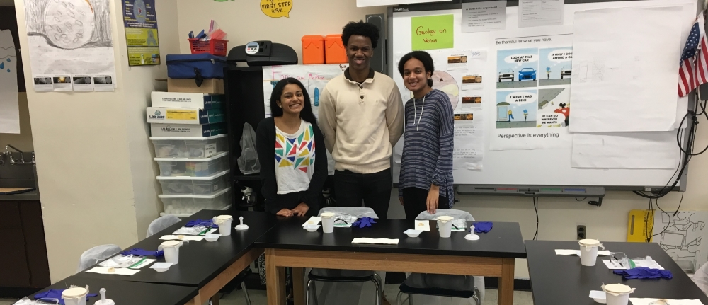Three students smiling behind desks in a science classroom